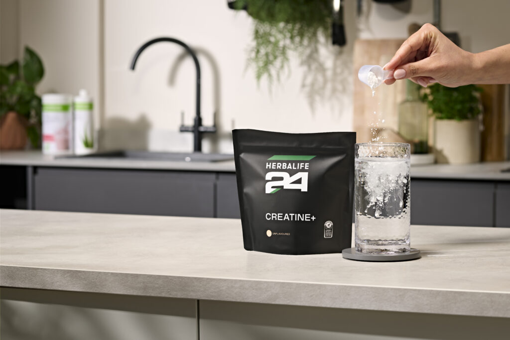 A person pouring a scoop of Herbalife24 Creatine+ powder into a glass of water on a kitchen counter.