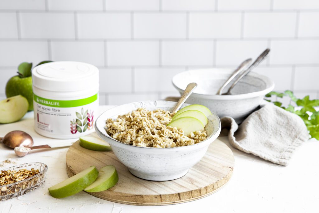 A tub of Herbalife Oat Apple Fibre Drink next to a healthy bowl of porridge topped with fresh apple slices; preparing a high-fibre breakfast in the UK
