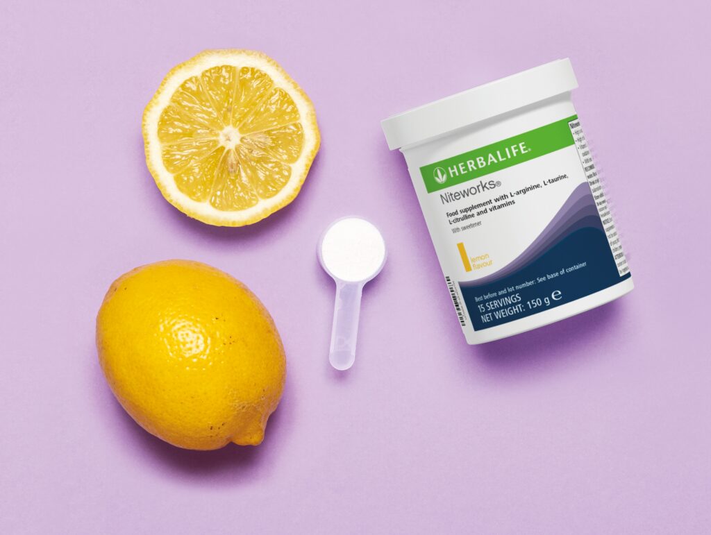 A flat-lay view of a Herbalife Niteworks lemon flavour tub next to a fresh lemon slice and a full measuring scoop on a purple background.