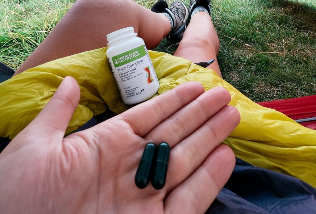 A close-up view of a person's open hand holding two dark green capsules, with a bottle of Herbalife Phyto Complete resting on a yellow jacket in an outdoor grassy setting.